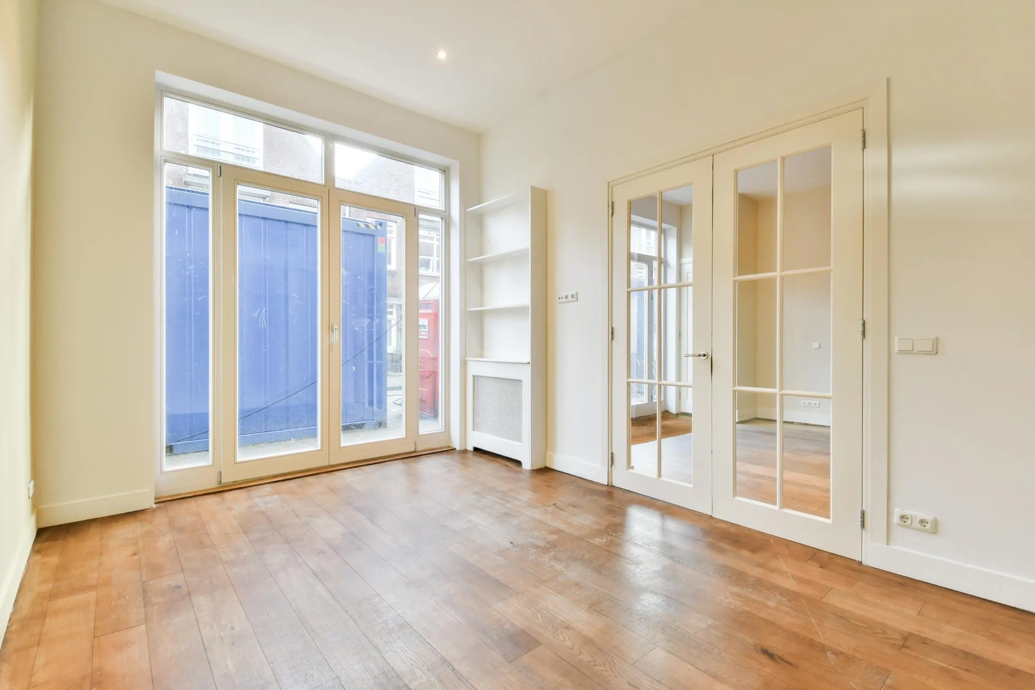 Lichte, lege kamer aan de Korte Schimmelstraat met houten vloer, glazen deuren en grote ramen met uitzicht op een blauwe container buiten.